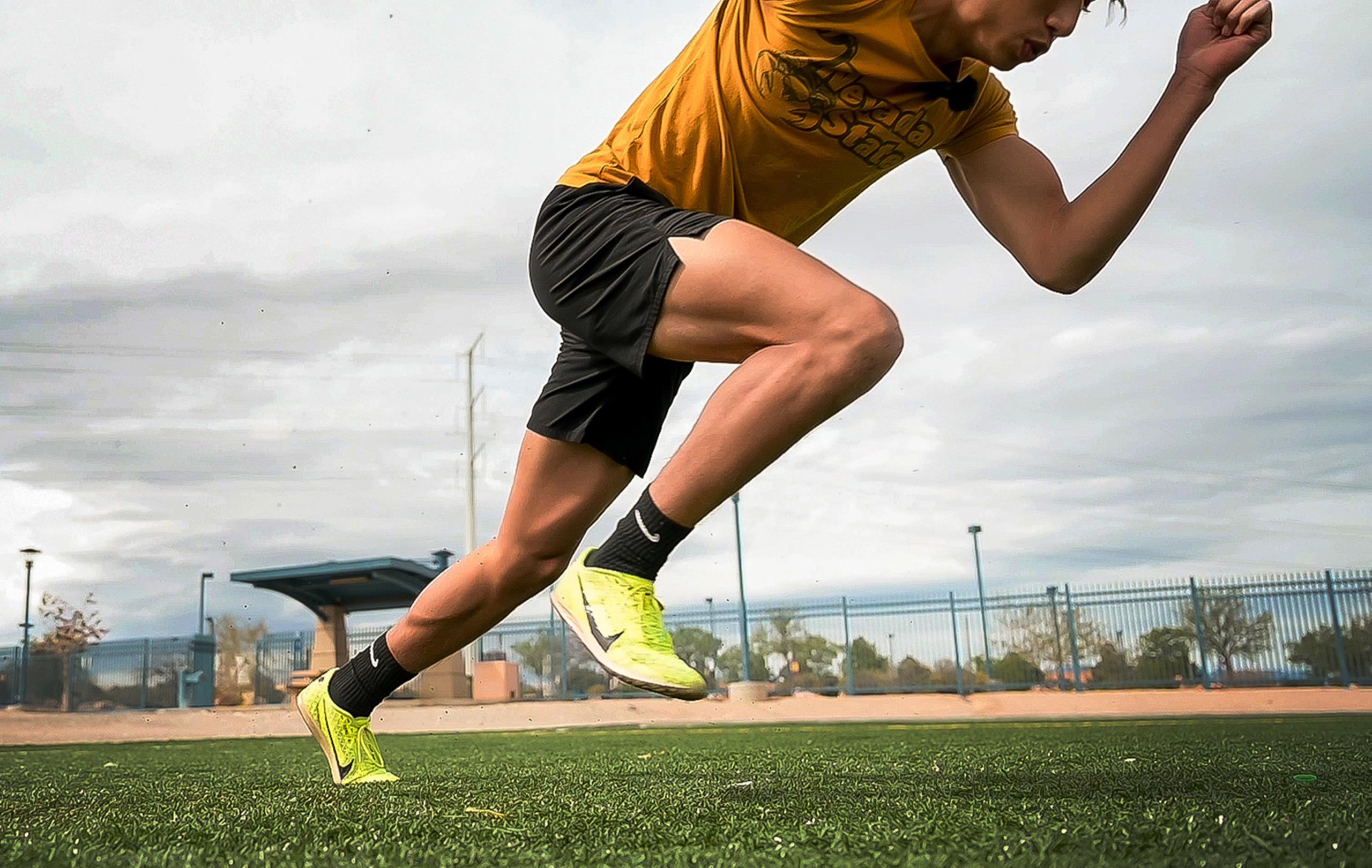 Male sprinter in Nevada State gold jersey and black shorts exploding off the starting line, showcasing explosive power in neon yellow running shoes against the track facility backdrop