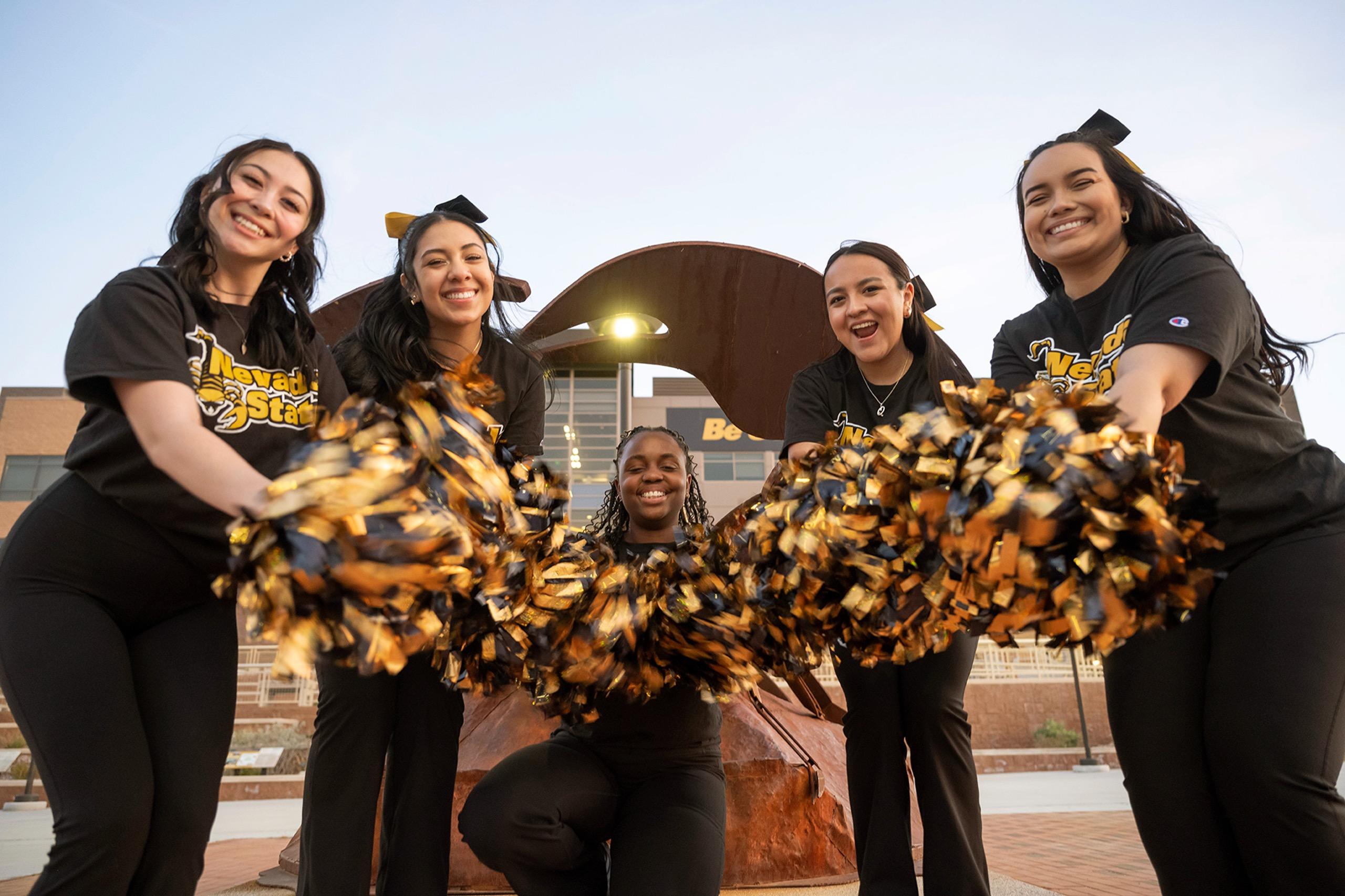 Group of five female Nevada State Dance Team student athletes smiling and shaking pom poms together, embodying teamwork and community